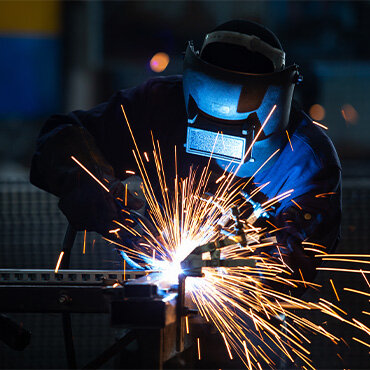 Welder wearing protective gear working with sparks flying during a welding process, joining metal components in a workshop.